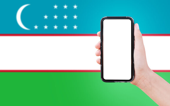 Close-up Of Male Hand Holding Smartphone With Blank On Screen, On Background Of Blurred Flag Of Uzbekistan.