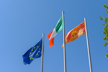 Italian, EU and the local flag of the Italian Veneto region waving in the wind. It is a beautiful sunny summer day, with a clear blue sky in the background.