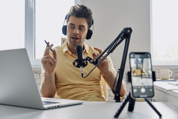 Young man in headphones using microphone and looking at laptop while recording podcast in studio