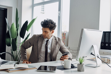 Naklejka premium Thoughtful young man holding smart phone while sitting at his working place in office