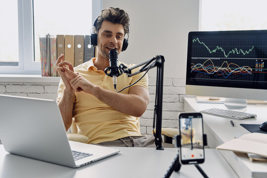 Young man in headphones using microphone and gesturing while recording his business podcast