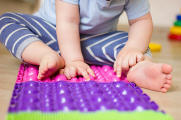 Toddler boy sitting on orthopedic massage mat and touch it by hands. Baby foot massage. Exercises for legs on carpet.