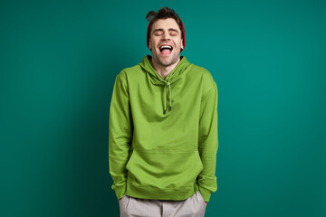 Happy young man laughing and keeping eyes closed while standing against green background