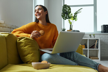Relaxed young woman using laptop and enjoying cookies while sitting on the couch at home