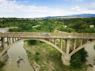 Fototapeta premium Car rides on an old arch bridge over a mountain river on a summer day, a side view from a drone