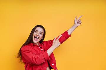 Happy young woman pointing away while standing against yellow background