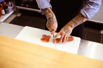Faceless view of tattooed chef cutting salami on cafe kitchen 