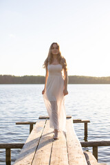 Beautiful Russian girl dressed in a white dress, walking along a wooden pier on the bank of a river or a lake