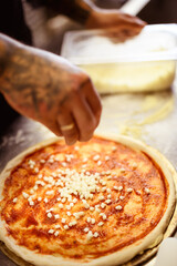Faceless shot of tattooed chef pouring cheese on raw pizza with sauce in cafe