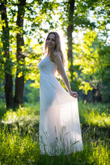 Beautiful young Russian girl dressed in a white dress, walks through the forest among the oaks in the rays of the sunset