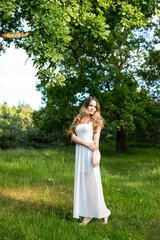 Beautiful young Russian girl dressed in a white dress, walks through the forest among the oaks in the rays of the sunset