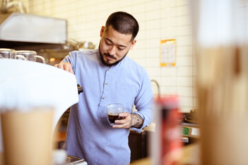 Tattooed asian cafe owner holding glass of coffee near coffee machine