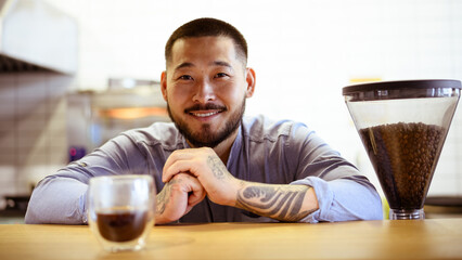 Tattooed asian barista smiling at camera near blurred glass of coffee in cafe