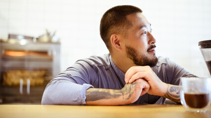 Obraz premium Asian barista looking away near blurred glass of americano in coffee shop 