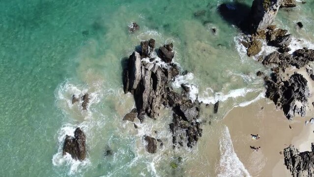 A cenital plane of a beautiful and  paradisiacal beach in which the waves are breaking onto the rocks and two children are playing in the shore with a board.