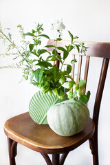 Autumn still life. A green bouquet in a vase of wormwood, mint, hops and phlox sprigs. A green pumpkin and a vase of flowers on a chair. Harvesting. Healthy lifestyle. Boho style