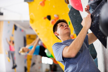 Young man climbing on rock-climbing wall during training in bouldering gym. - obrazy, fototapety, plakaty