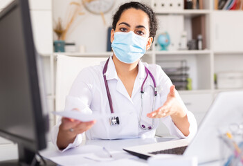 Portrait of confident woman doctor in face mask explaining prescription to patient in office in...