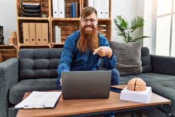 Redhead man with long beard using laptop working at psychology clinic smiling happy pointing with...