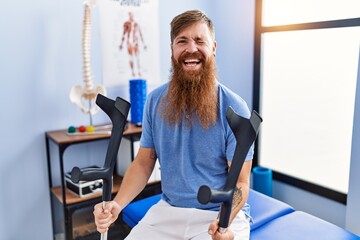 Redhead man with long beard holding crutches at rehabilitation clinic winking looking at the camera...