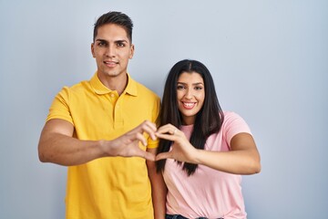 Young couple standing over isolated background smiling in love doing heart symbol shape with hands. romantic concept.