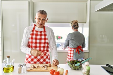 Middle age caucasian couple cooking healthy salad smiling happy pointing with hand and finger