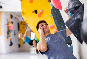 European young man having telephone conversation while grabbing ledges of artificial climbing wall in bouldering centre. © JackF