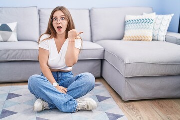 Obraz premium Young caucasian woman sitting on the floor at the living room surprised pointing with hand finger to the side, open mouth amazed expression.