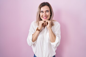 Young beautiful woman standing over pink background laughing nervous and excited with hands on chin looking to the side