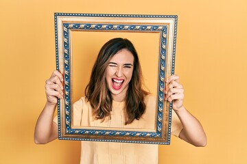 Young hispanic girl holding empty frame winking looking at the camera with sexy expression, cheerful and happy face.