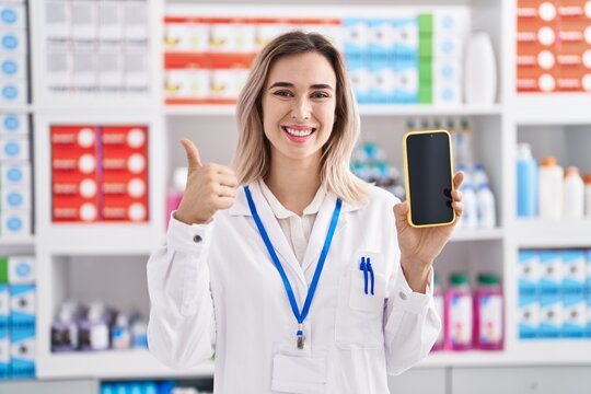 Young beautiful woman working at pharmacy drugstore showing smartphone screen smiling happy and positive, thumb up doing excellent and approval sign