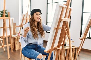 Young hispanic artist woman painting on canvas at art studio sticking tongue out happy with funny expression. emotion concept.