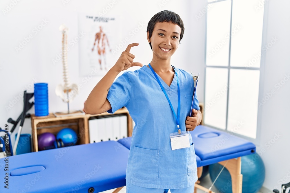 Sticker Young hispanic woman with short hair working at pain recovery clinic smiling and confident gesturing with hand doing small size sign with fingers looking and the camera. measure concept.