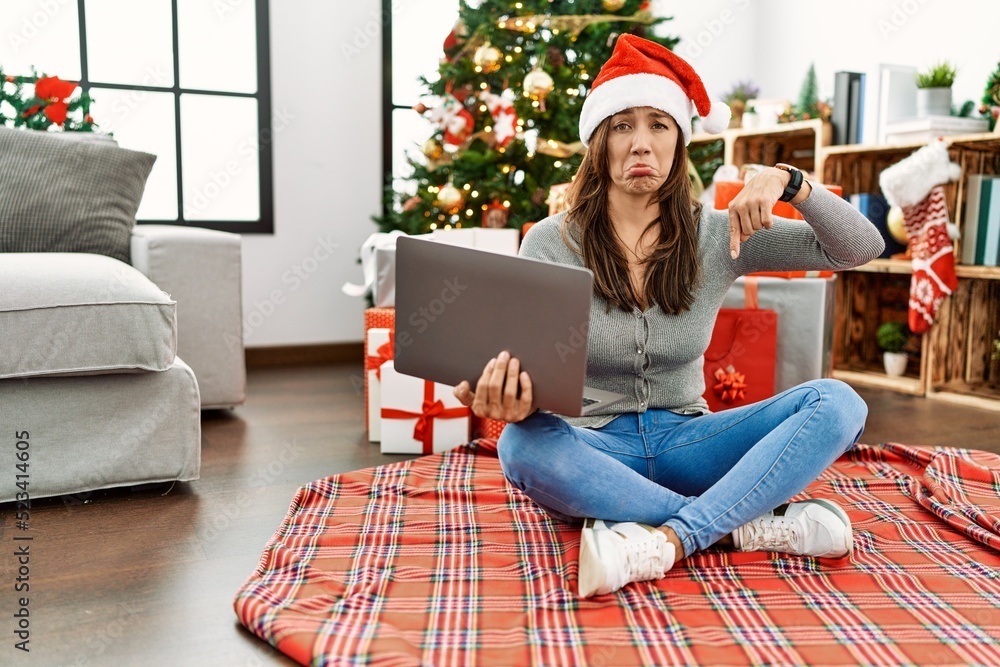 Sticker Young latin woman using laptop sitting by christmas tree pointing down looking sad and upset, indicating direction with fingers, unhappy and depressed.