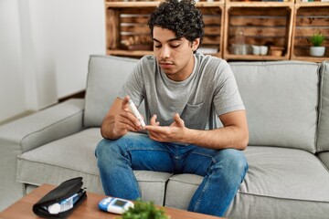 Young hispanic diabetic man measuring glucose sitting on the sofa at home.