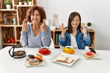 Family of mother and down syndrome daughter sitting at home eating breakfast gesturing finger crossed smiling with hope and eyes closed. luck and superstitious concept.