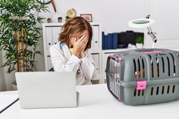 Middle age veterinarian woman working at pet clinic with sad expression covering face with hands...