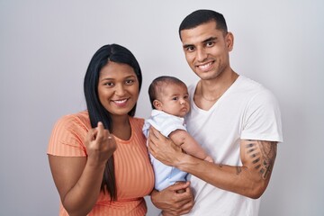 Young hispanic couple with baby standing together over isolated background beckoning come here gesture with hand inviting welcoming happy and smiling