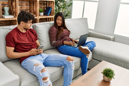 Young Latin Couple With Serious Expression Using Laptop And Smartphone At Home.