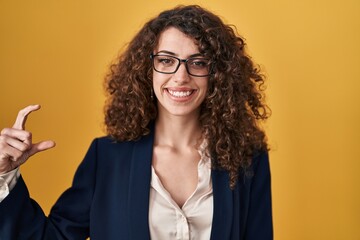 Hispanic woman with curly hair standing over yellow background smiling and confident gesturing with hand doing small size sign with fingers looking and the camera. measure concept.