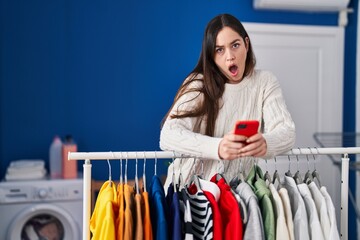 Young brunette woman waiting for laundry using smartphone in shock face, looking skeptical and...