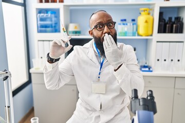 African american man working at scientist laboratory holding syringe covering mouth with hand, shocked and afraid for mistake. surprised expression