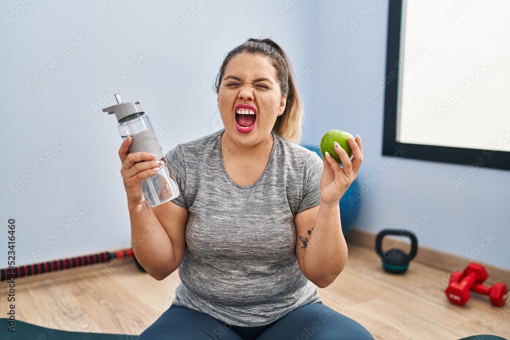 Poster Young hispanic woman holding green apple and bottle of water angry and mad screaming frustrated and furious, shouting with anger. rage and aggressive concept.