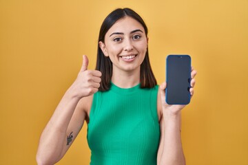 Hispanic girl holding smartphone showing screen smiling happy and positive, thumb up doing excellent and approval sign