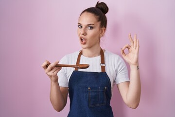 Young hispanic girl wearing professional cook apron holding wood spoon in shock face, looking skeptical and sarcastic, surprised with open mouth