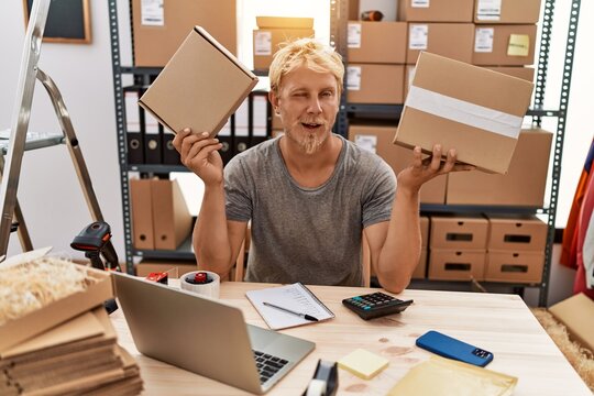Young blond man holding packages working at online shop winking looking at the camera with sexy expression, cheerful and happy face.