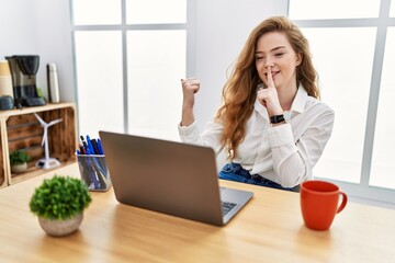 Young caucasian woman working at the office using computer laptop asking to be quiet with finger on lips pointing with hand to the side. silence and secret concept.