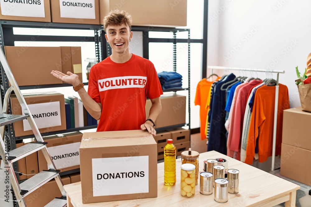 Wall mural Young caucasian man volunteer holding donations box pointing aside with hands open palms showing copy space, presenting advertisement smiling excited happy