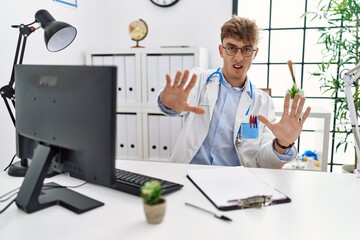 Young caucasian doctor man working at the clinic afraid and terrified with fear expression stop gesture with hands, shouting in shock. panic concept.