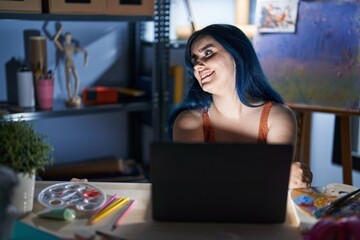 Fototapeta na wymiar Young modern girl with blue hair sitting at art studio with laptop at night looking away to side with smile on face, natural expression. laughing confident.